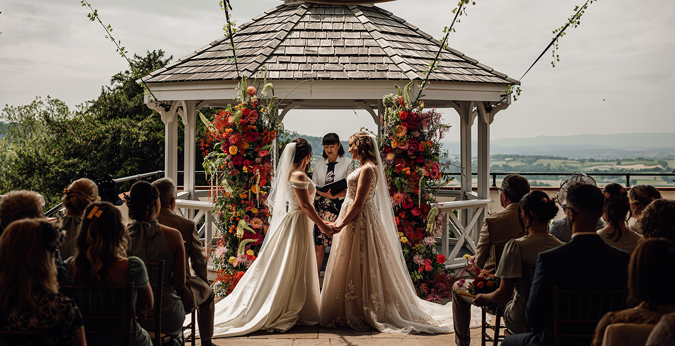 A wedding under a flower-adorned gazebo with two brides, officiant, and guests overlooking scenery.