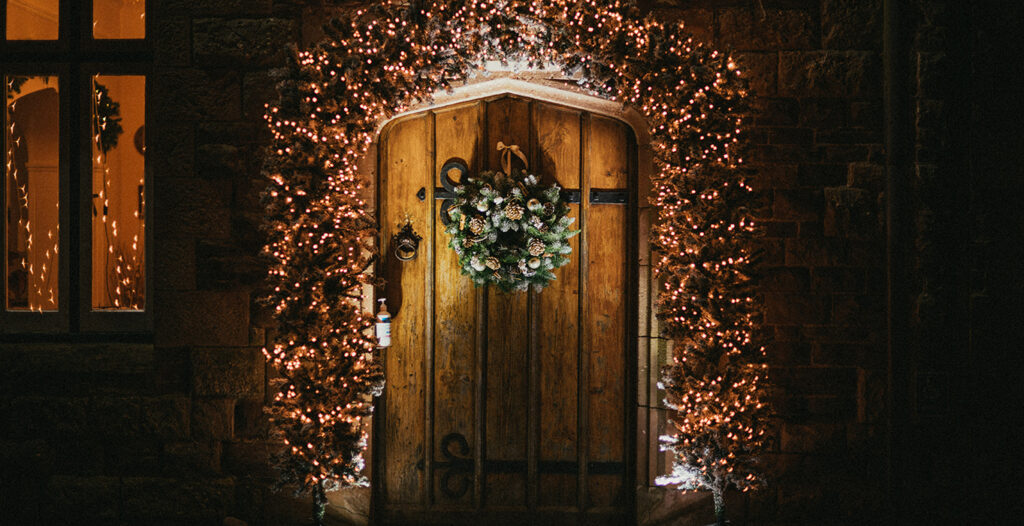 Festive wreath on wooden door with lights, stone wall, and window reflecting warm glow.