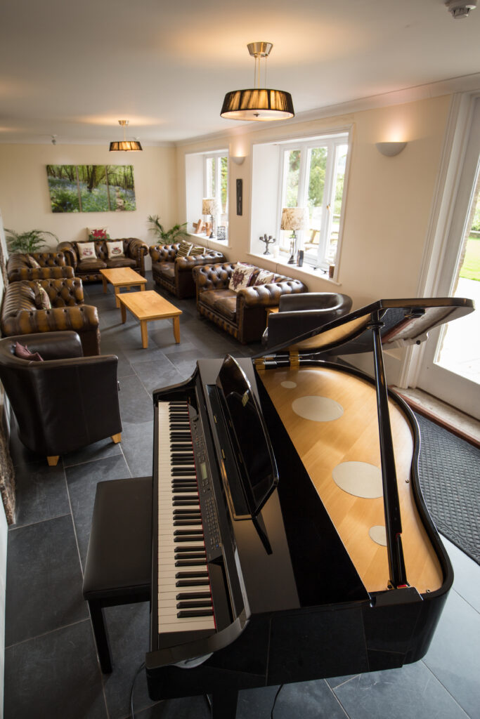 Living room with brown leather sofas, piano, wooden tables, large windows, and nature painting.