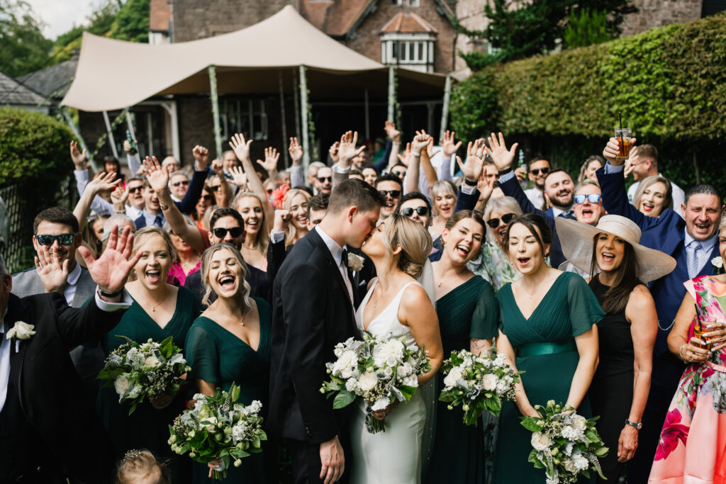 Caer Llan Bride and groom kissing outdoors with bridesmaids in green dresses and guests celebrating.