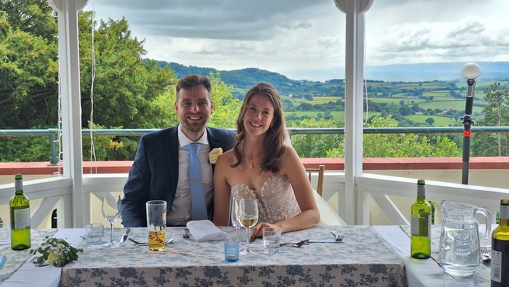 Caer Llan Newlywed couple at a decorated table on a covered terrace overlooking a rural landscape.